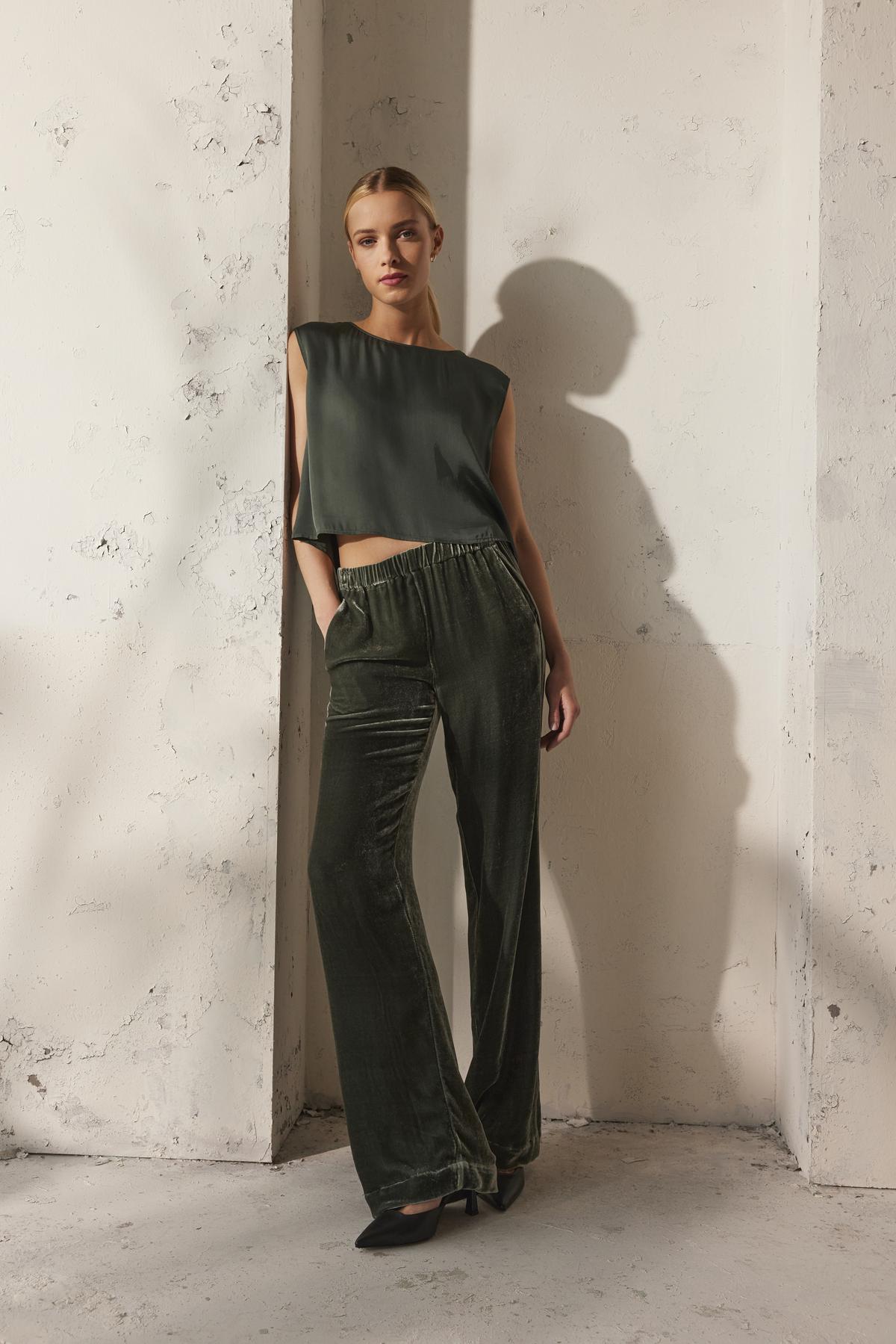 A person wearing a sleeveless top in a deep green hue paired with the SORINE SILK VELVET PANT from Velvet by Graham & Spencer is standing against a textured off-white wall.-38246701957313