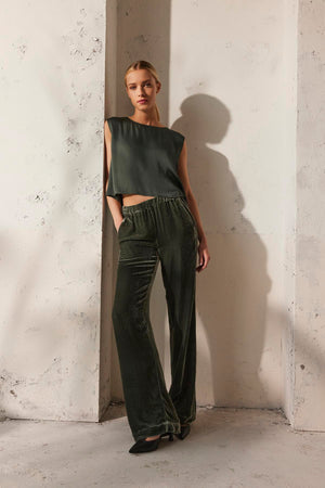 A person wearing a sleeveless top in a deep green hue paired with the SORINE SILK VELVET PANT from Velvet by Graham & Spencer is standing against a textured off-white wall.