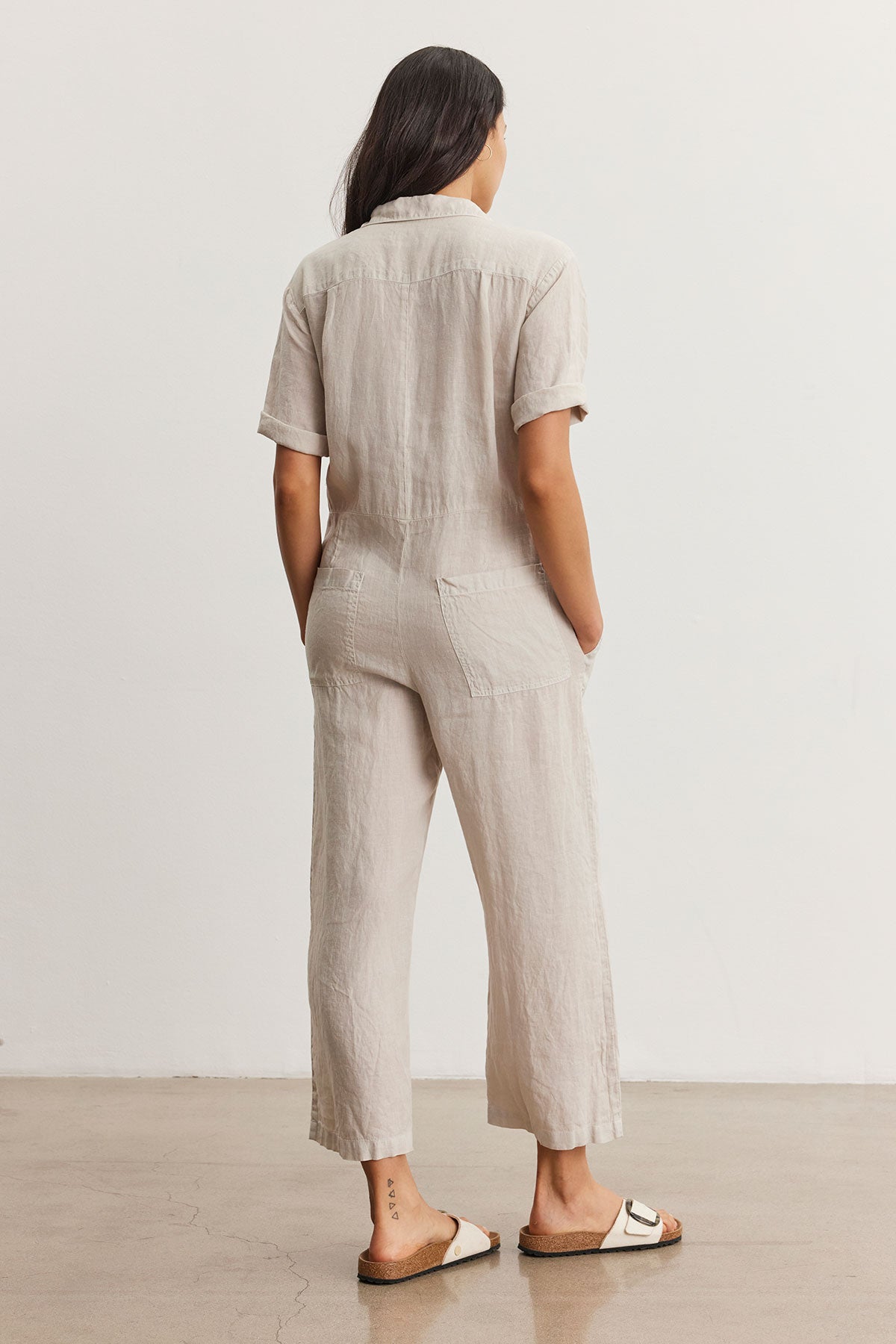 Woman standing facing away, wearing a beige short-sleeved Velvet by Graham & Spencer DELILAH LINEN JUMPSUIT and white sandals, in a minimalist room.-36998726615233