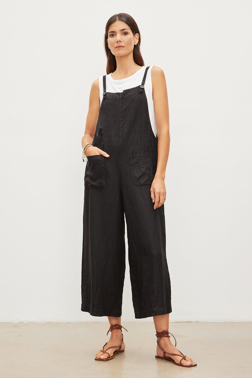 Woman standing in a white studio wearing black Velvet by Graham & Spencer ISABEL LINEN JUMPERS with patch pockets and a white shirt, paired with brown sandals, hands in pockets, looking at the camera.