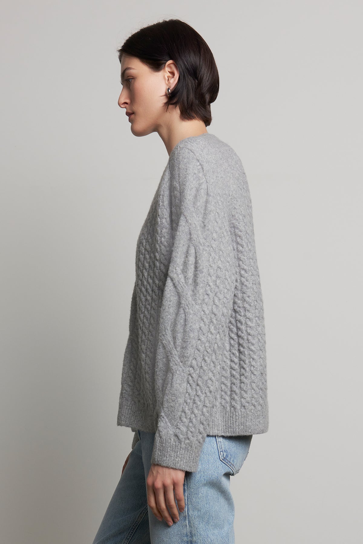 A person wearing the WILSHIRE SWEATER, a gray wool-yak blend cable-knit piece by Velvet by Jenny Graham, paired with jeans, stands against a plain background, facing left.-38318578434241