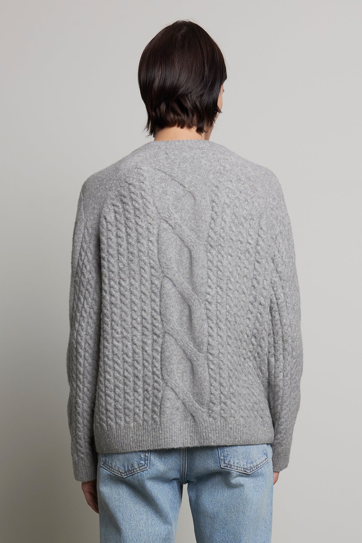   A person is photographed from the back against a neutral background wearing the WILSHIRE SWEATER, a gray wool-yak blend cable-knit piece by Velvet by Jenny Graham, paired with light blue jeans. 