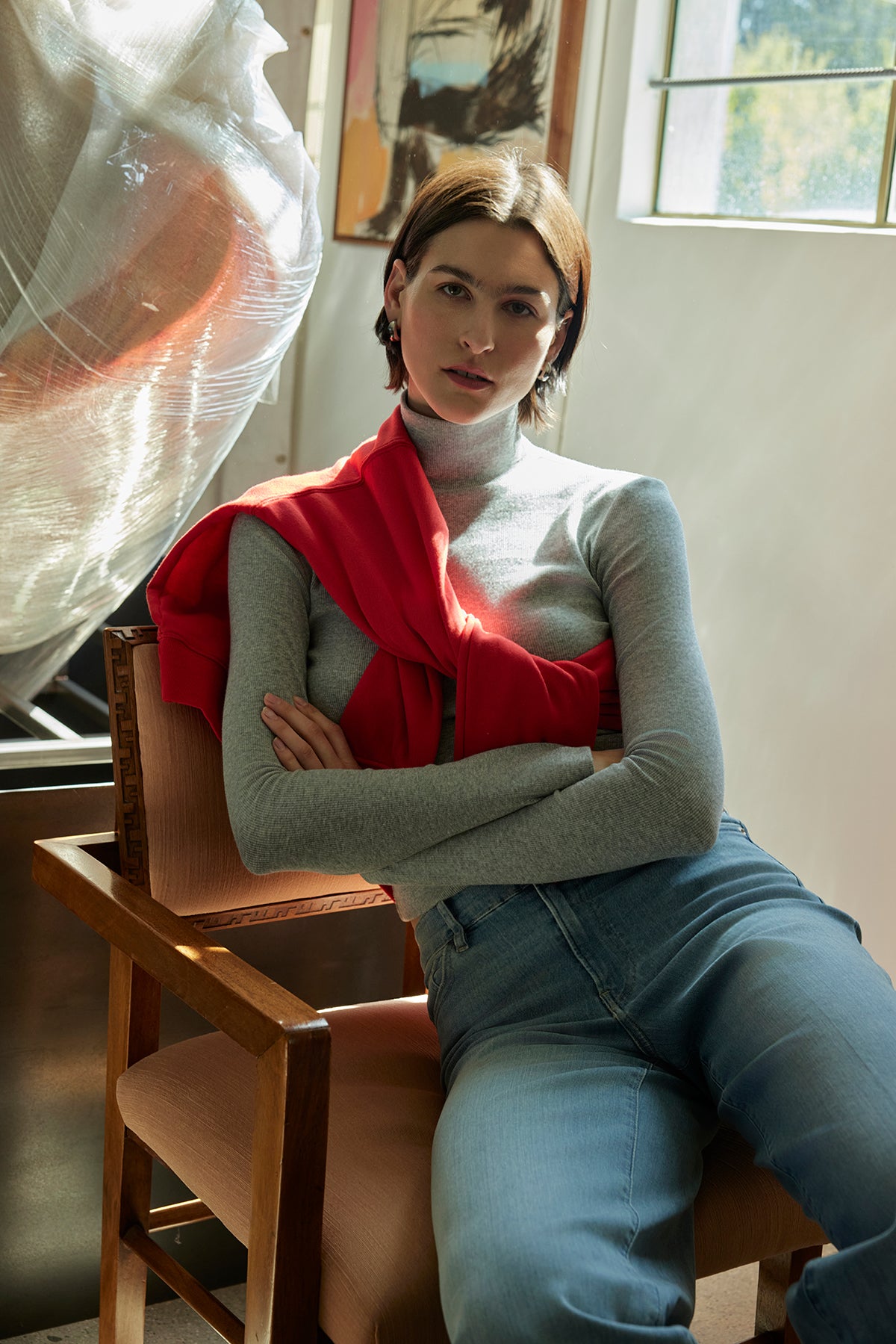   A person sits on a chair, dressed in the HELMS TEE by Velvet by Jenny Graham and jeans, with a red sweater draped over their shoulders, in a sunlit room adorned with abstract art. 