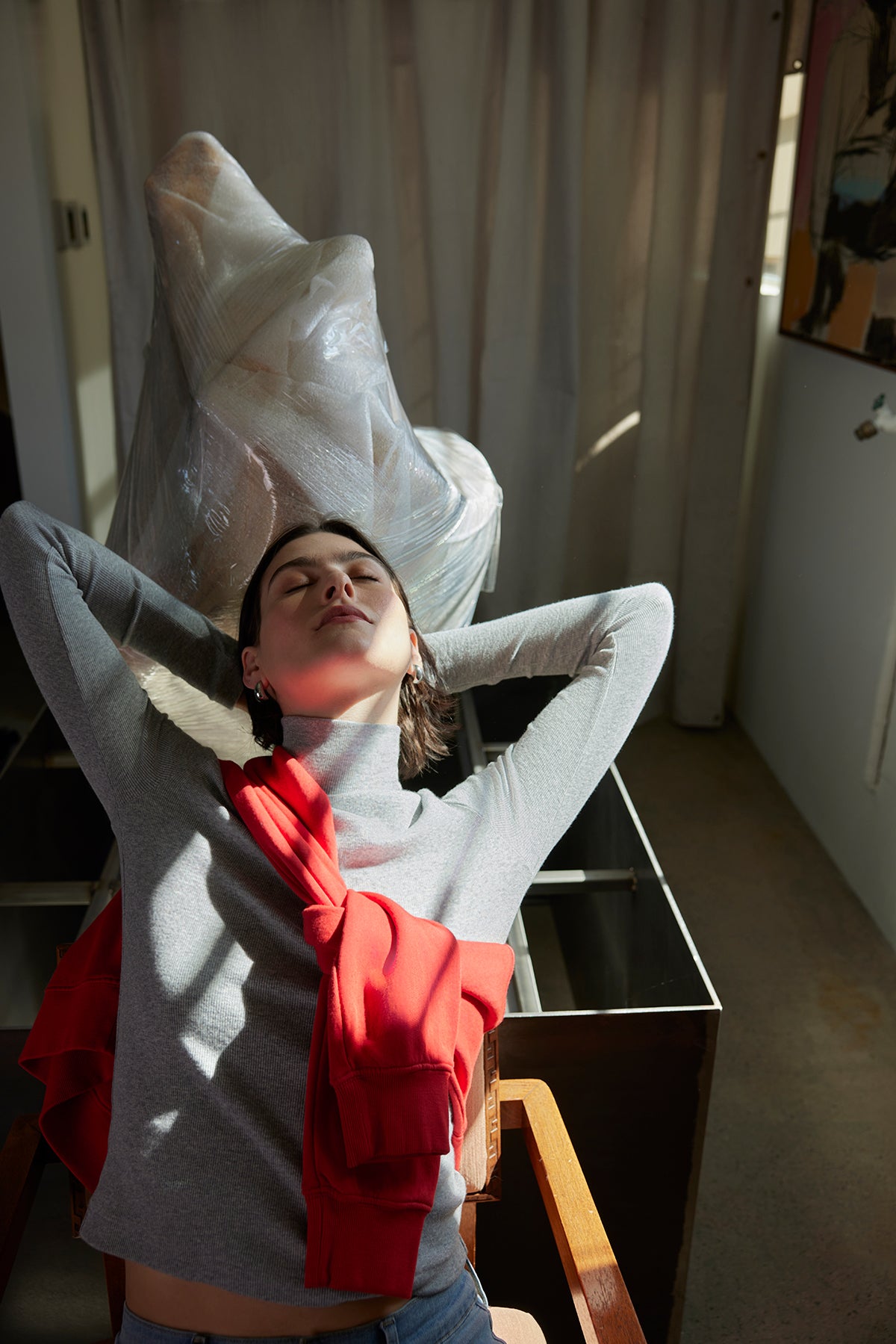   A person wearing the HELMS TEE by Velvet by Jenny Graham in a gray ribbed style leans back with eyes closed in a chair, near a large wrapped item and a partially curtained window, with a red garment draped over them. 