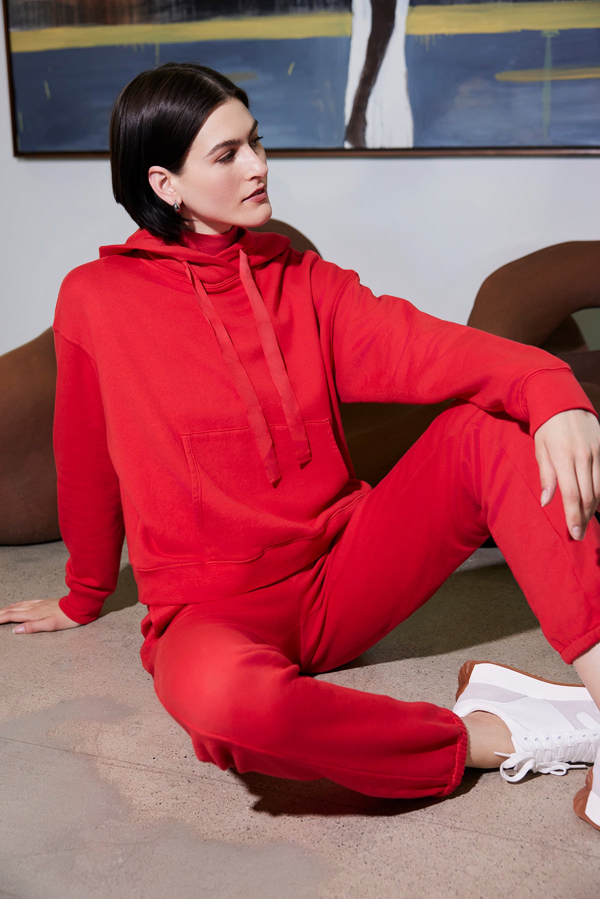   A person in an oversized OJAI HOODIE by Velvet by Jenny Graham and matching pants sits on the floor with one knee bent. White sneakers complement the look, while art and a brown chair adorn the background, enhancing this relaxed scene. 