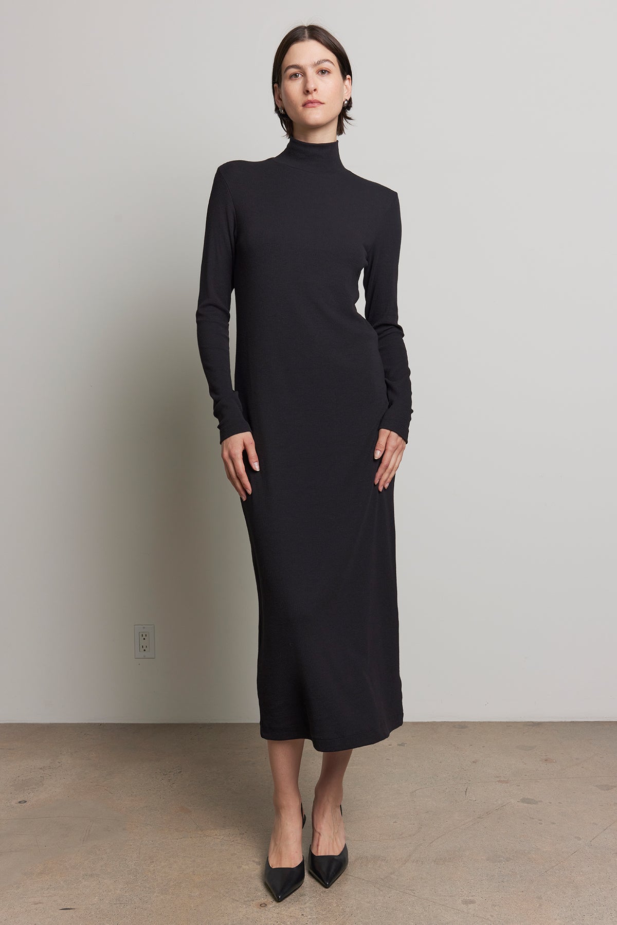   A person wearing the AGOURA DRESS by Velvet by Jenny Graham, styled with black pointed shoes, stands against a plain background. 