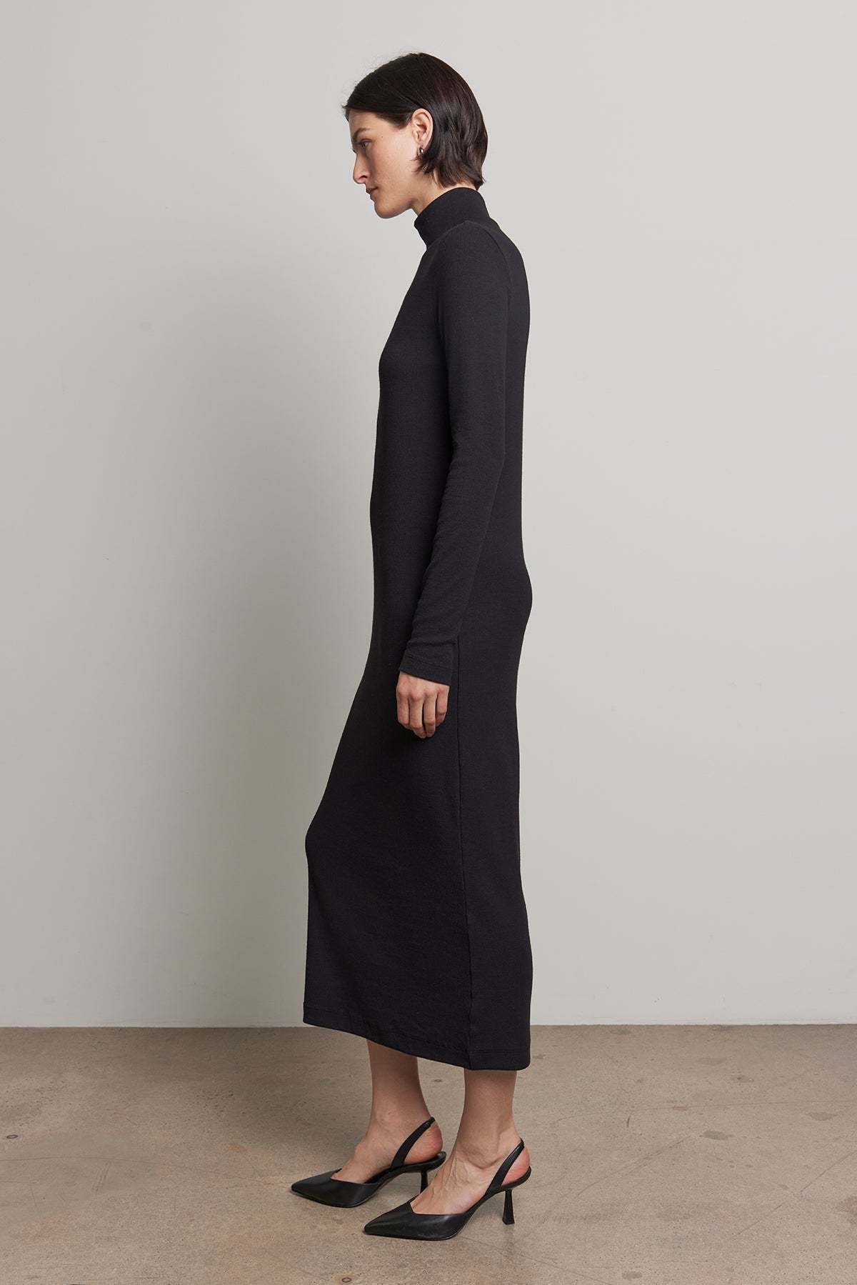   A person wearing the AGOURA DRESS by Velvet by Jenny Graham, a sleek black midi dress with a fitted silhouette, paired with black heels, stands in profile against a plain background. 