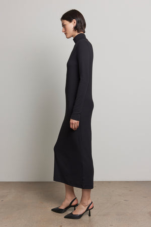 A person wearing the AGOURA DRESS by Velvet by Jenny Graham, a sleek black midi dress with a fitted silhouette, paired with black heels, stands in profile against a plain background.