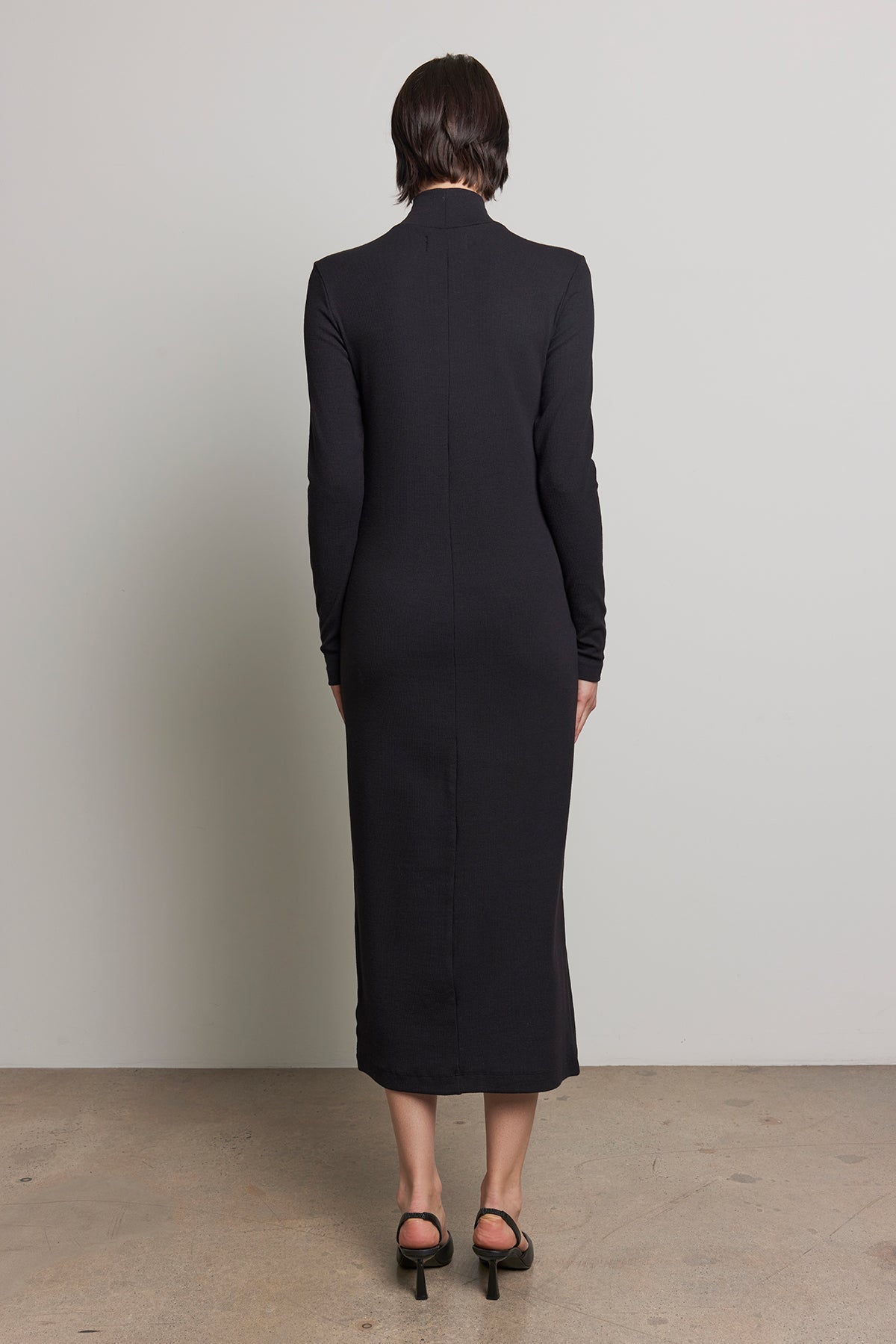   With their short hair turned away from the camera, the individual is wearing the AGOURA DRESS, a sleek long-sleeved black midi dress by Velvet by Jenny Graham, paired with black heels. 