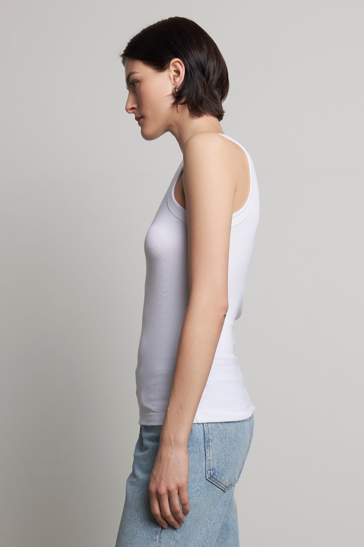   The individual is wearing the CRUZ TANK TOP from Velvet by Jenny Graham, paired with stretch blue jeans, standing sideways against a plain background and perfectly embodying the essence of a capsule wardrobe. 