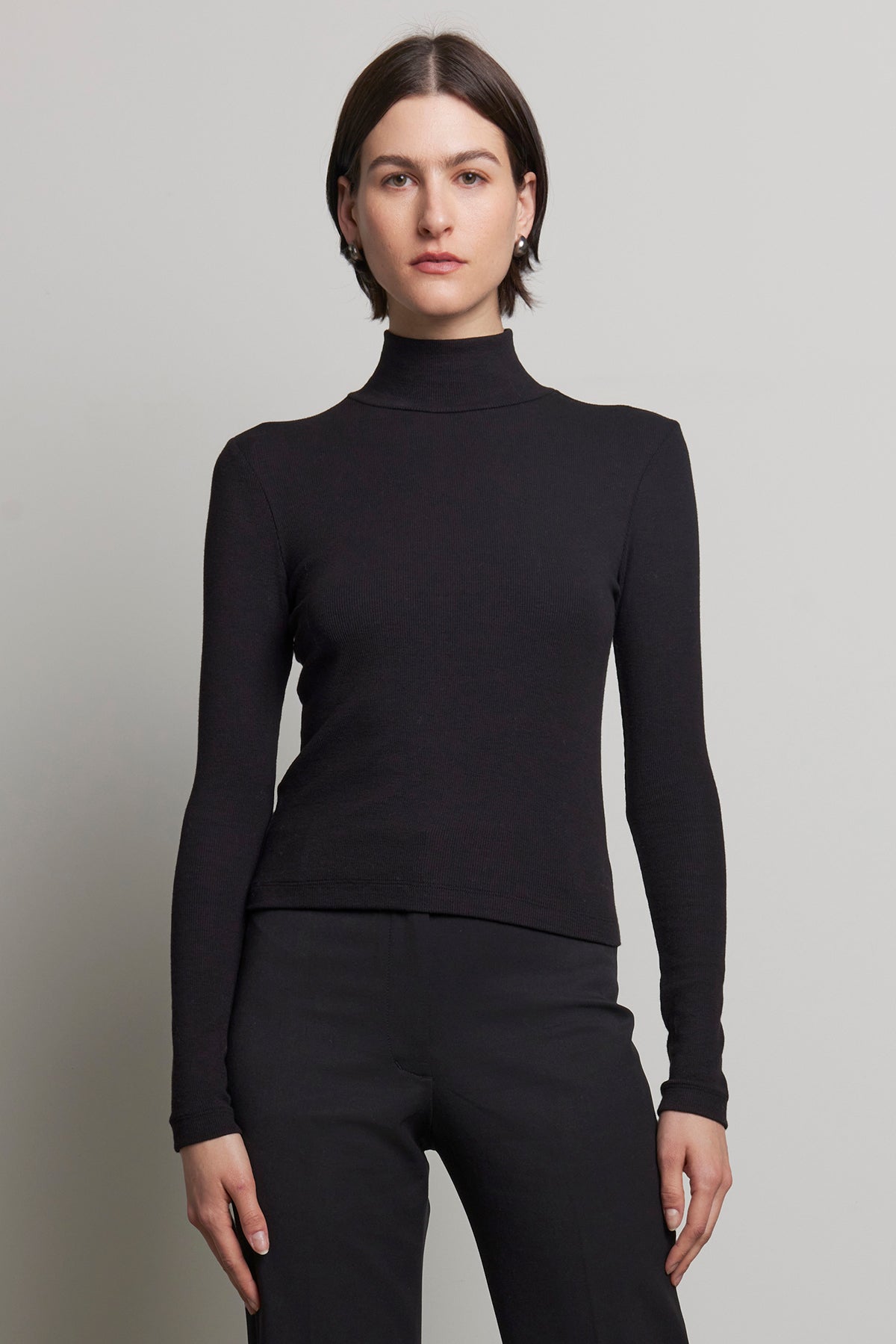 A person stands against a neutral background, dressed in the sleek HELMS TEE by Velvet by Jenny Graham, featuring a black long-sleeve turtleneck with a subtle ribbed texture and matching pants.-38318596882625