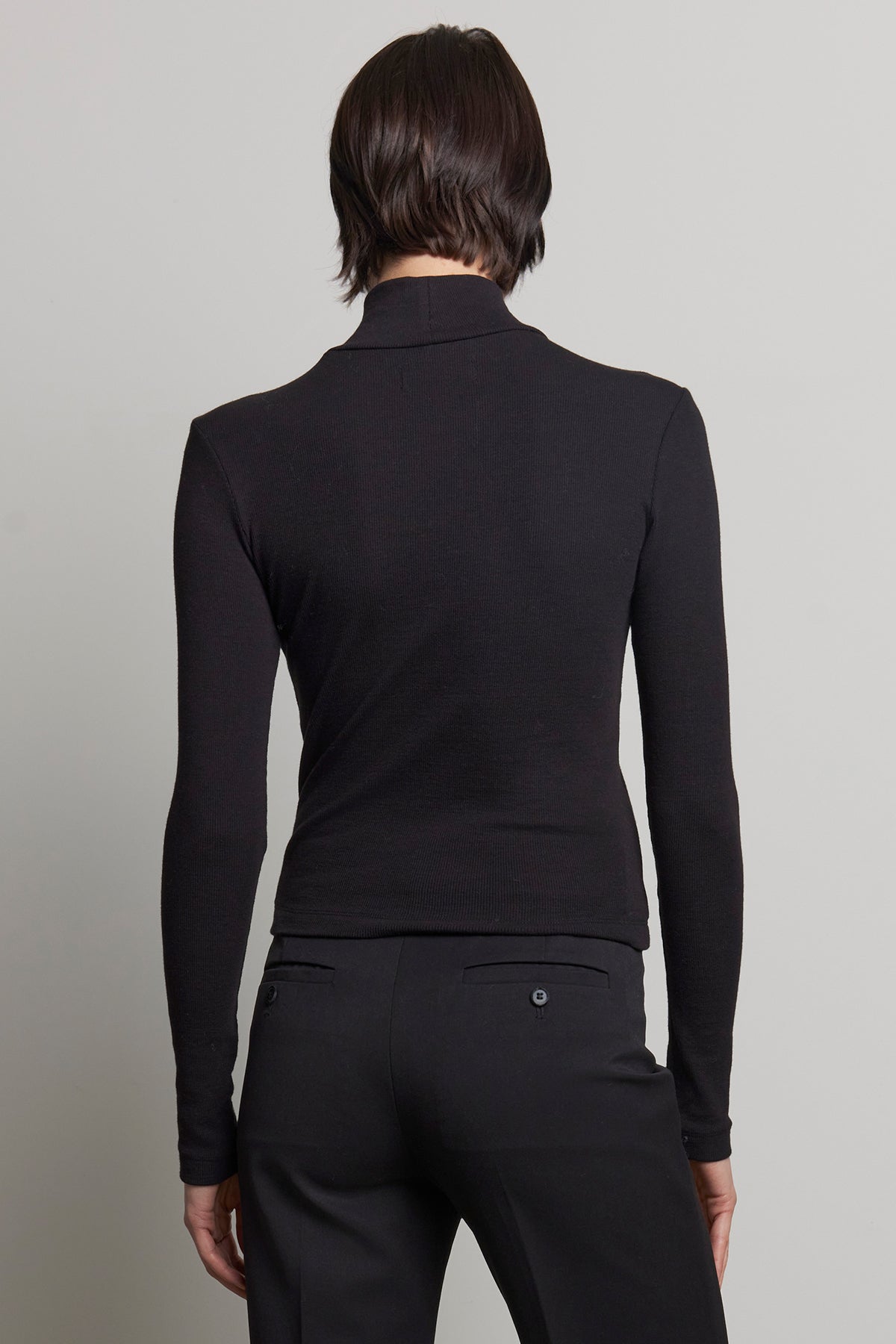   A person with short dark hair is wearing the HELMS TEE, a long-sleeved black mock neck top by Velvet by Jenny Graham, along with black ribbed textured pants, seen from the back against a light gray background. 