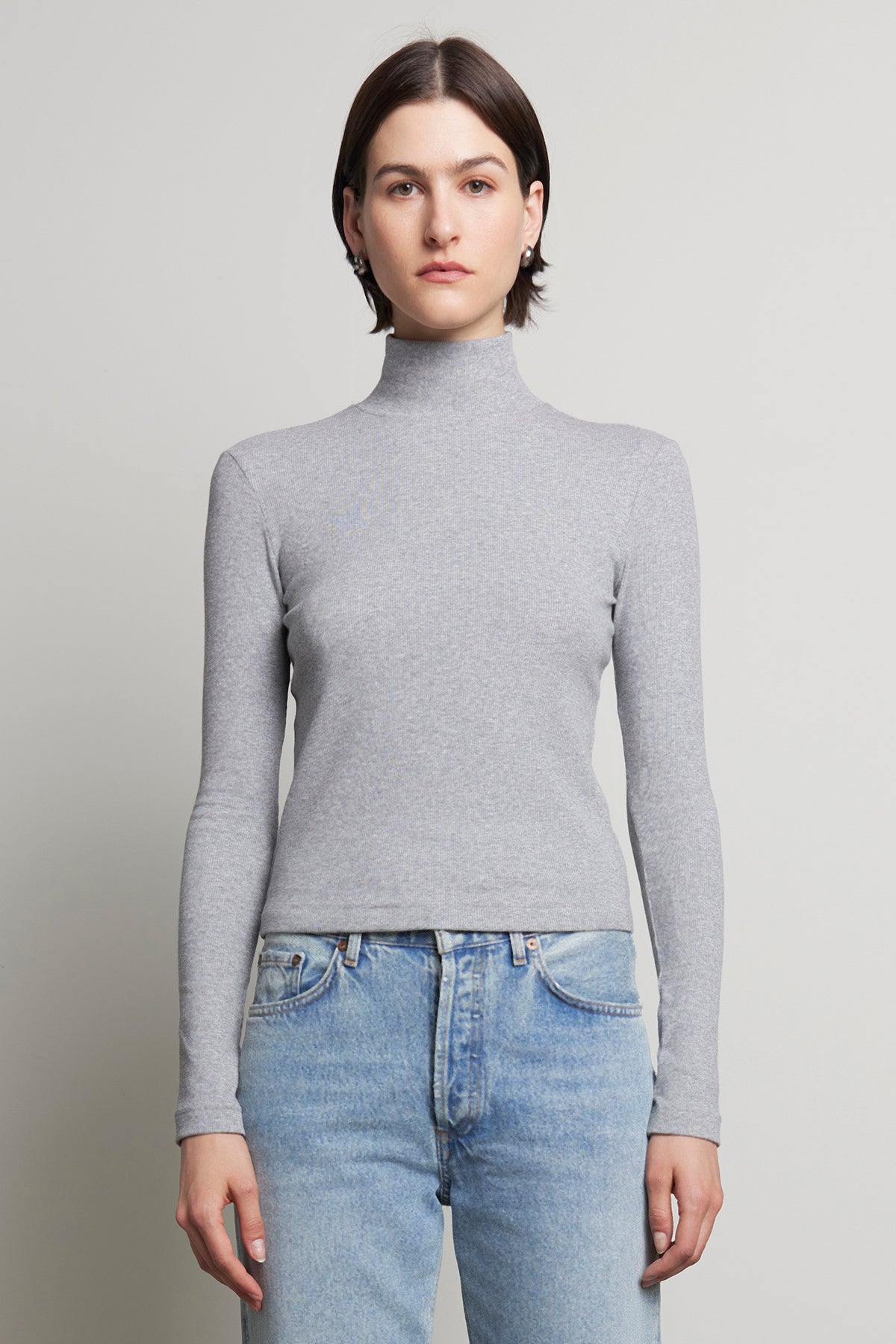   A person wearing the HELMS TEE by Velvet by Jenny Graham and blue jeans stands against a plain background, with the ribbed texture of their mock neck gray turtleneck adding subtle depth to the minimalist scene. 