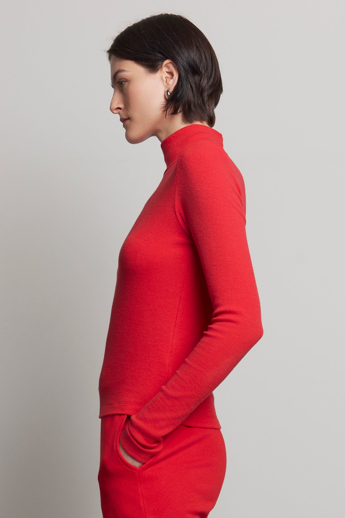 A person in profile is wearing the HELMS TEE, a red turtleneck with a subtle ribbed texture from Velvet by Jenny Graham, paired with pants and standing against a plain background.-38318597046465