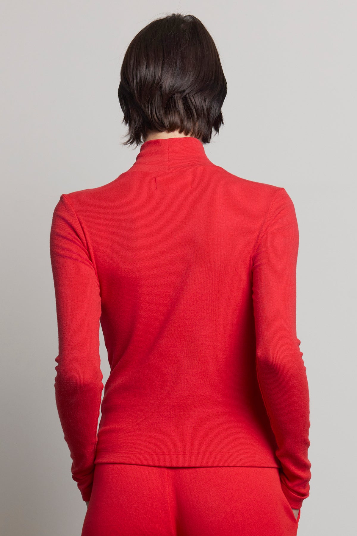 A person with short brown hair is wearing the HELMS TEE, a long-sleeved, red turtleneck sweater from Velvet by Jenny Graham, featuring a ribbed texture. They are facing away from the camera against a plain gray background.-38318597112001