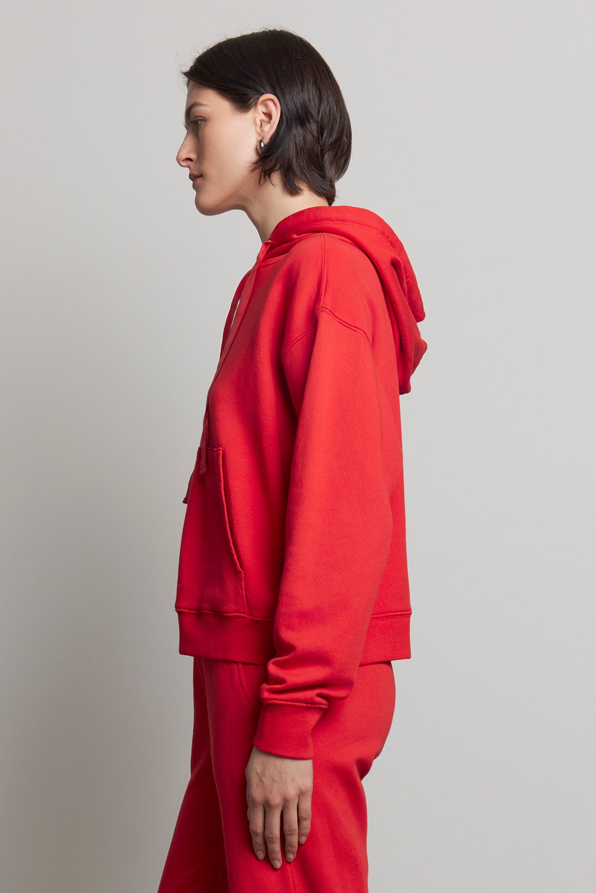 A person wearing an oversized OJAI HOODIE in red from the brand Velvet by Jenny Graham, paired with organic cotton pants, stands in profile against a plain background.-38318612676801