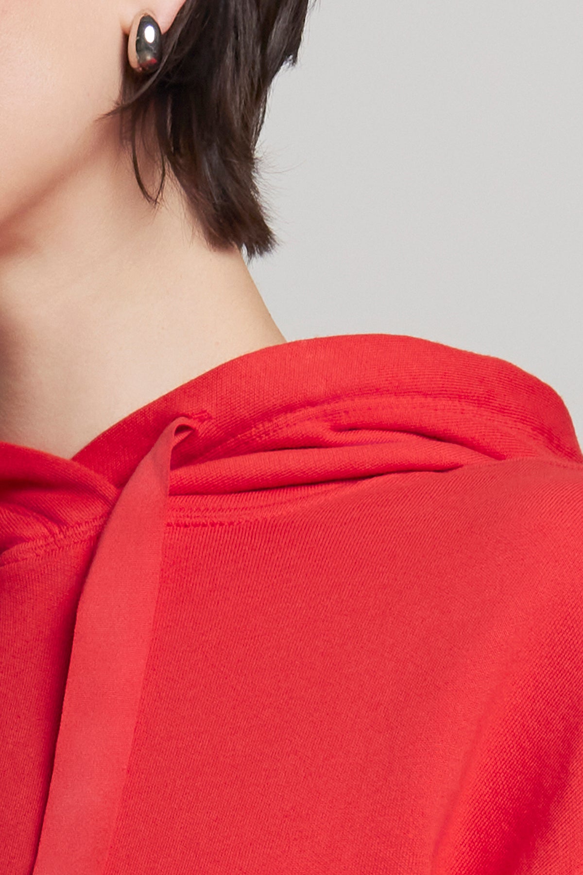   Close-up of a person wearing the OJAI HOODIE by Velvet by Jenny Graham, featuring classic red organic cotton, with a single stud earring and short dark hair visible. 