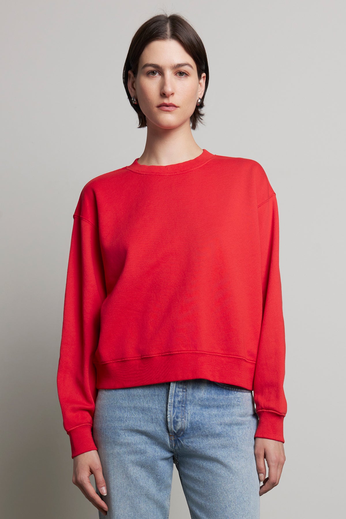 A person with short hair, wearing a red YNEZ SWEATSHIRT by Velvet by Jenny Graham and blue jeans, stands against a plain background.-38318624506049