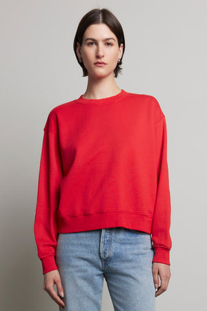 A person with short hair, wearing a red YNEZ SWEATSHIRT by Velvet by Jenny Graham and blue jeans, stands against a plain background.