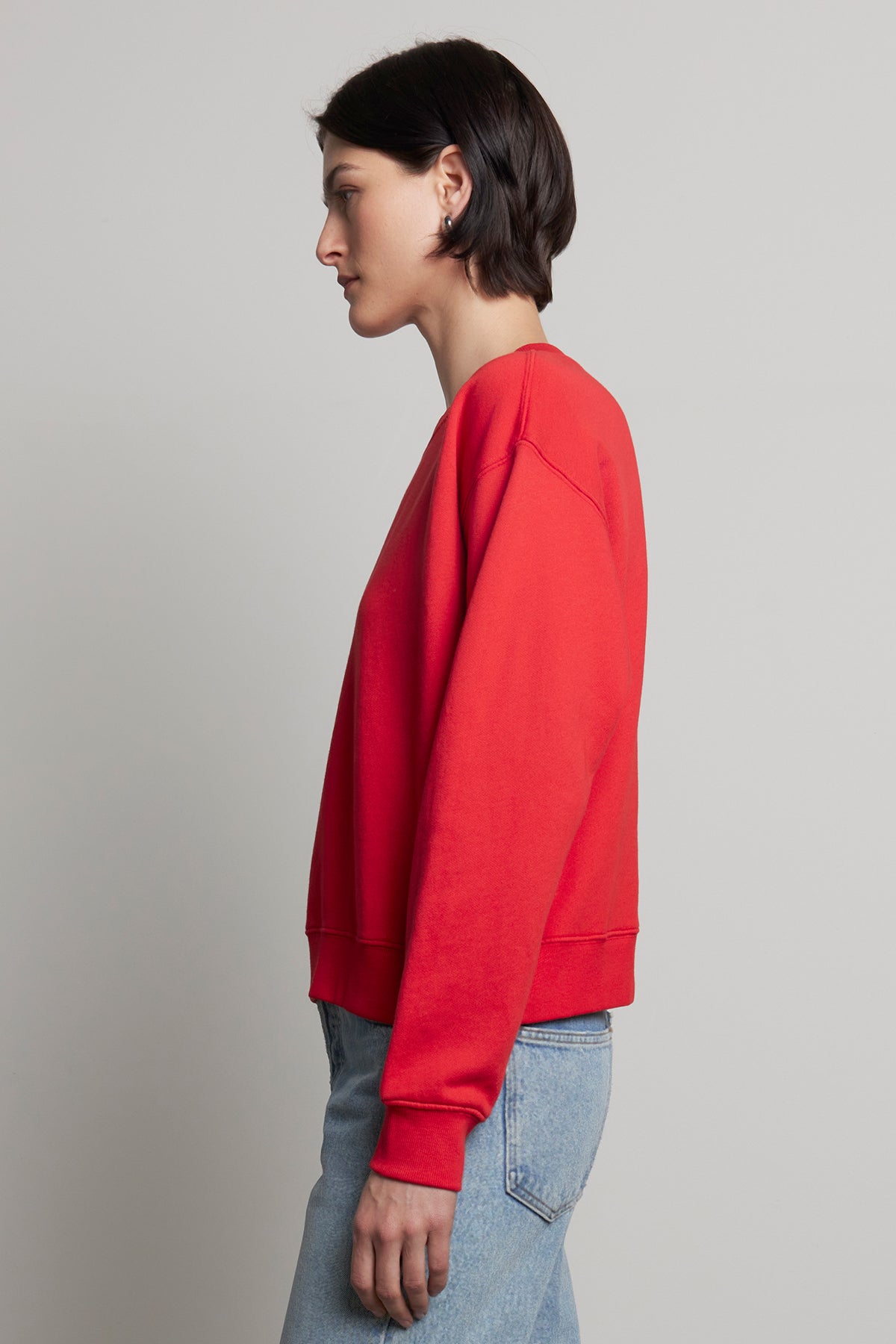 A person in a YNEZ sweatshirt by Velvet by Jenny Graham and blue jeans, standing in profile against a plain background, highlighting the elegance of sustainable fashion.-38318624571585