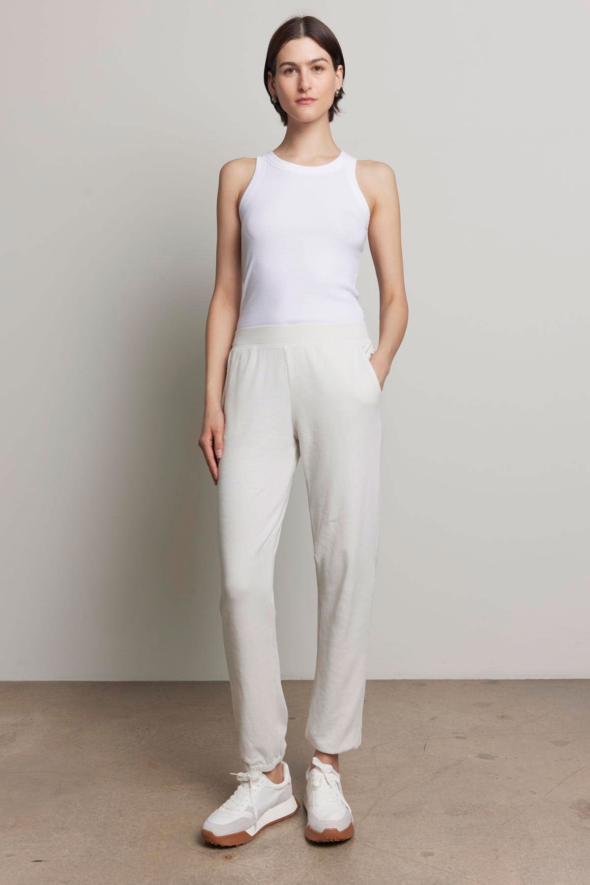   A person is standing against a plain background, dressed in a white tank top and the ZUMA SWEATPANT from Velvet by Jenny Graham, featuring off-white organic cotton material with an elastic waist and pockets, paired with white sneakers, looking at the camera with a neutral expression. 