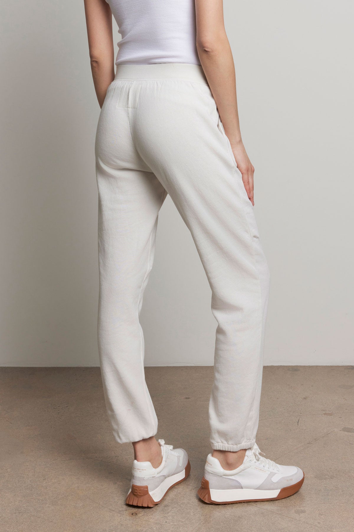   Someone in a white tank top and Velvet by Jenny Graham's ZUMA SWEATPANT stands facing away, highlighting the back of their outfit with its elastic waist. They are wearing white sneakers with tan soles. 