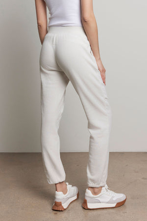 Someone in a white tank top and Velvet by Jenny Graham's ZUMA SWEATPANT stands facing away, highlighting the back of their outfit with its elastic waist. They are wearing white sneakers with tan soles.