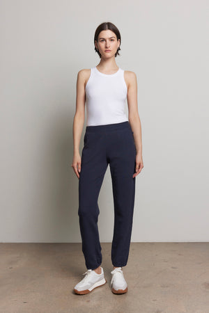 A person is posing against a gray wall dressed in a white sleeveless organic cotton top, ZUMA SWEATPANT by Velvet by Jenny Graham, and white sneakers.