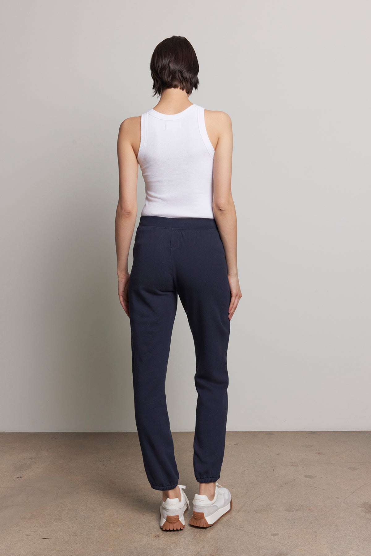   A person wearing a white tank top and ZUMA SWEATPANT from Velvet by Jenny Graham, made from organic cotton, is standing with their back to the camera against a plain background. 