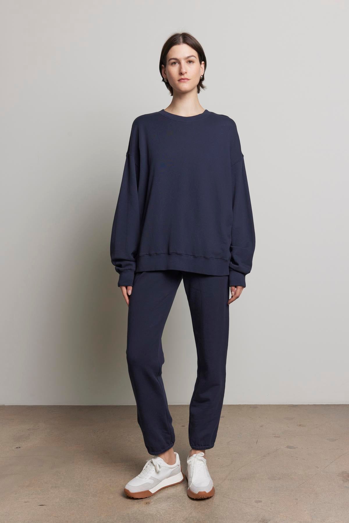   A person stands against a plain background wearing the YNEZ SWEATSHIRT in navy blue from Velvet by Jenny Graham, paired with matching pants and white sneakers. 