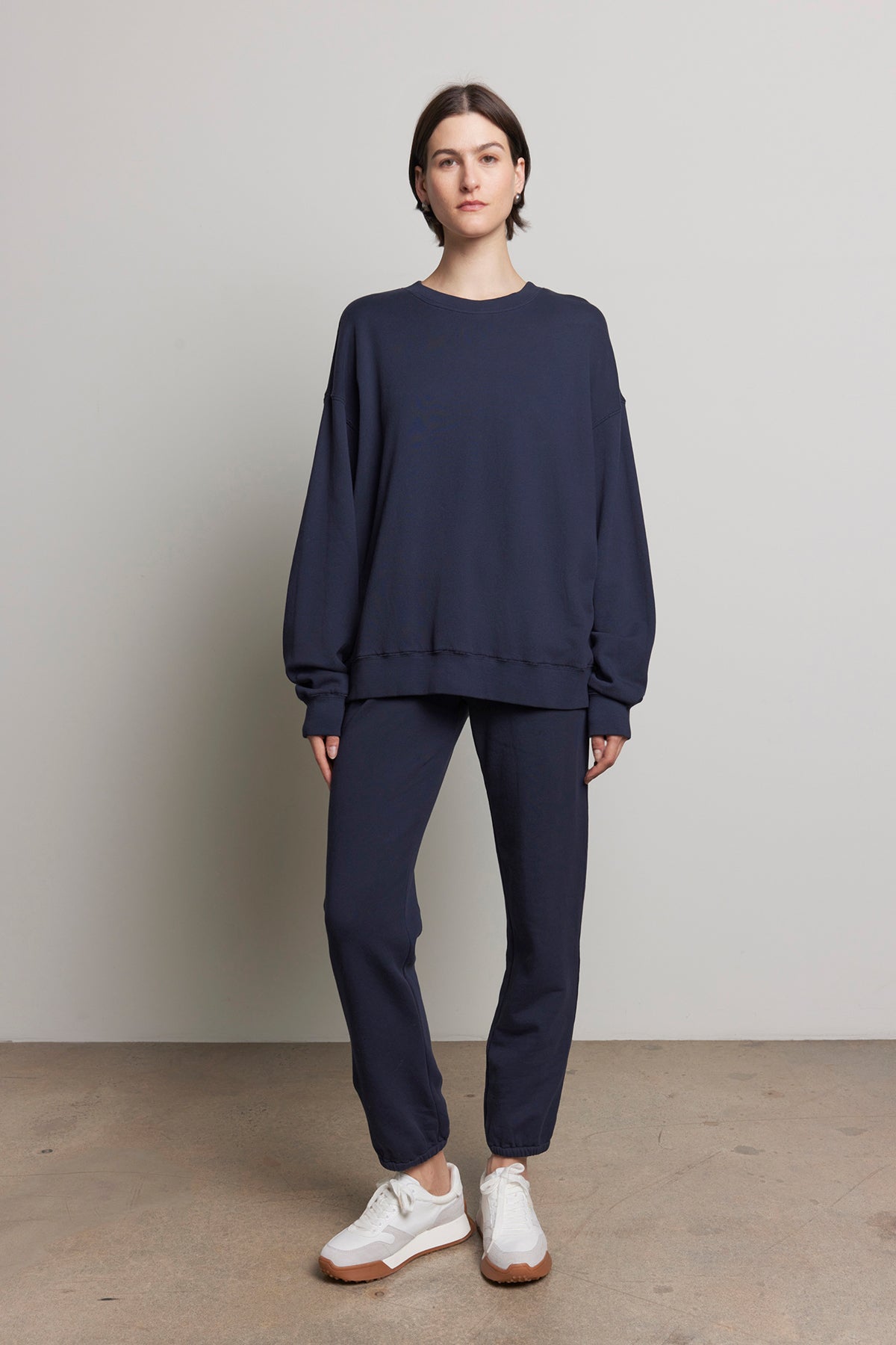 A person wearing a navy sweatshirt and ZUMA SWEATPANT joggers from Velvet by Jenny Graham, made from organic cotton and paired with white sneakers, stands against a plain light gray wall.-38318659633345