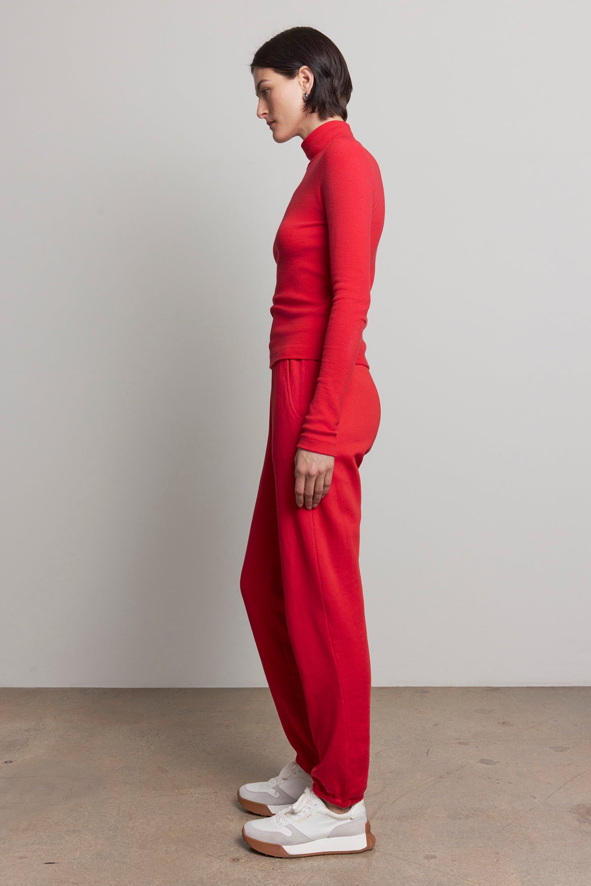   Dressed in a vibrant red turtleneck and Velvet by Jenny Graham's ZUMA SWEATPANT, an individual showcases sustainable fashion while wearing white sneakers against a plain background. 