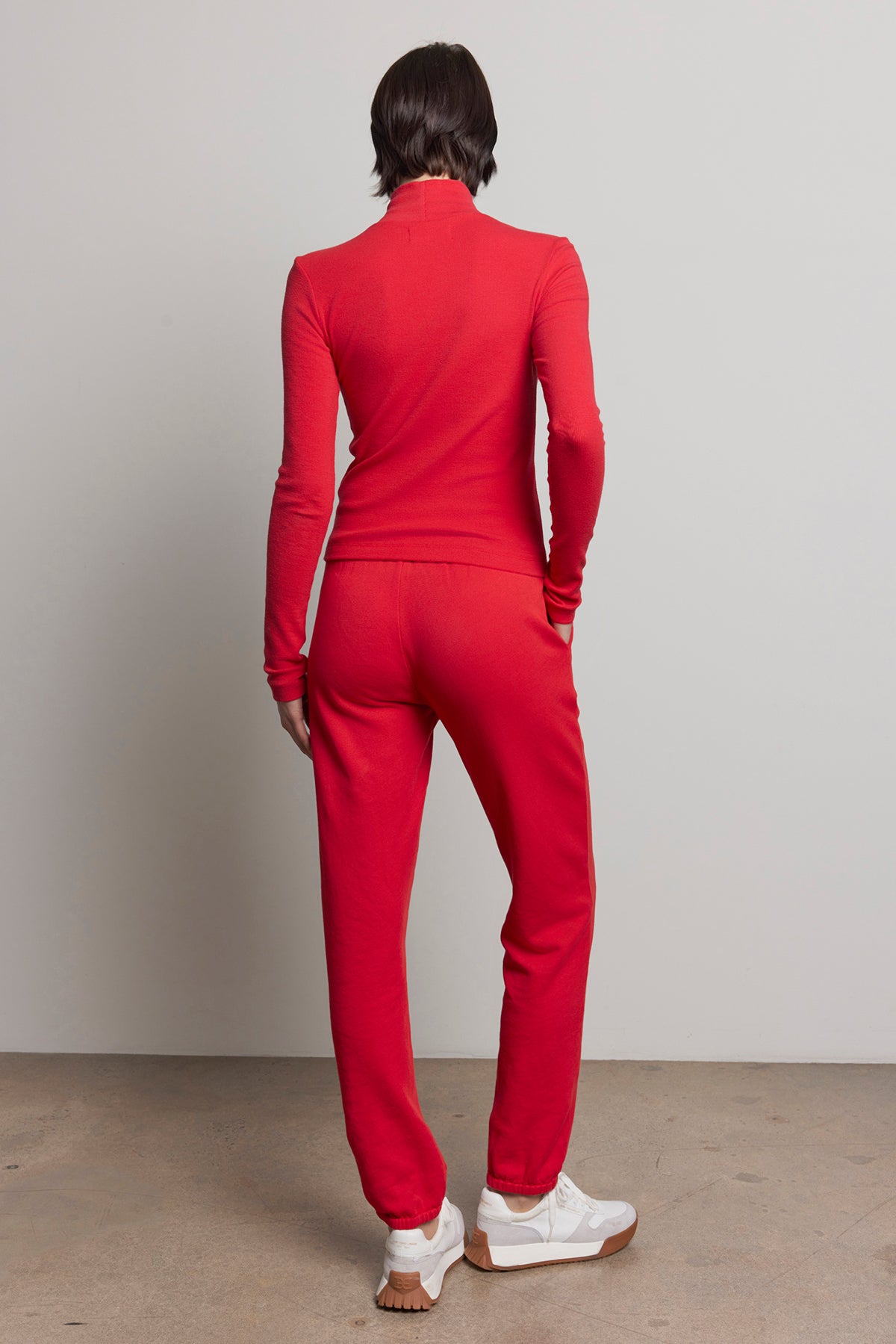   A person wearing a red long-sleeve top and the ZUMA SWEATPANT by Velvet by Jenny Graham stands on a concrete floor, facing away. White sneakers provide contrast to the outfit, embodying sustainable fashion. 