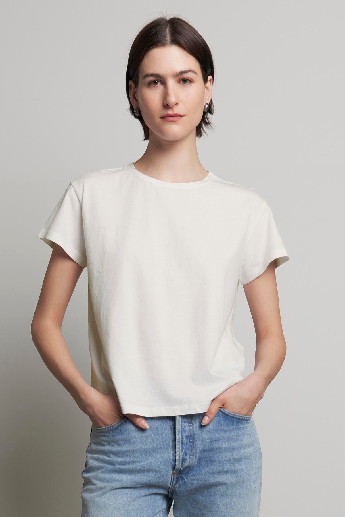 A person with short, dark hair is wearing the TOPANGA TEE by Velvet by Jenny Graham, made of organic cotton, paired with relaxed-fit classic blue jeans, standing with hands in pockets against a plain backdrop.-37817548177601