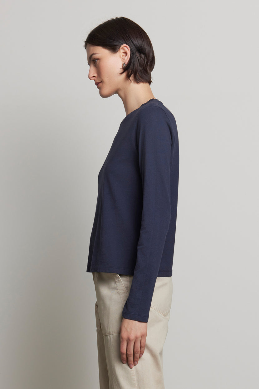 A person with short dark hair stands sideways against a plain background, wearing Velvet by Jenny Graham's VICENTE TEE, a relaxed-fit long sleeve navy tee made of organic cotton, paired with beige pants.