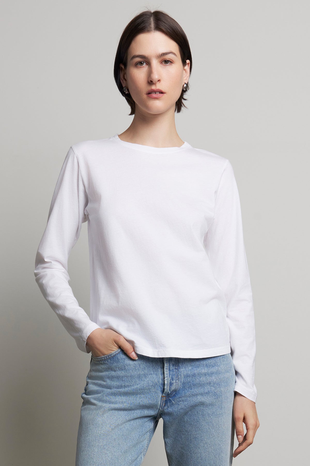   A person wearing the VICENTE TEE by Velvet by Jenny Graham, a relaxed-fit white long-sleeve shirt made from organic cotton, and blue jeans stands against a plain background. 