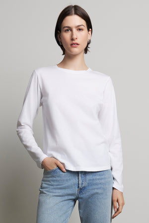 A person wearing the VICENTE TEE by Velvet by Jenny Graham, a relaxed-fit white long-sleeve shirt made from organic cotton, and blue jeans stands against a plain background.