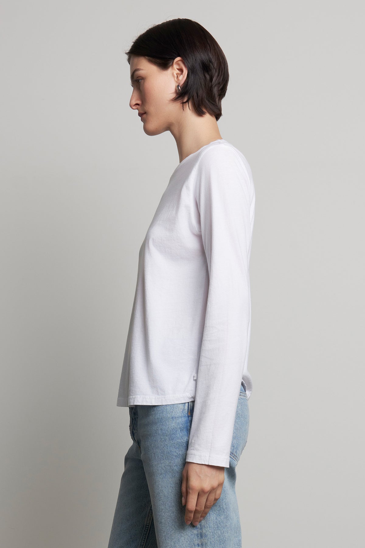 A person wearing the VICENTE TEE, a white long-sleeve organic cotton shirt from Velvet by Jenny Graham, and relaxed-fit blue jeans stands sideways.-38866409980097