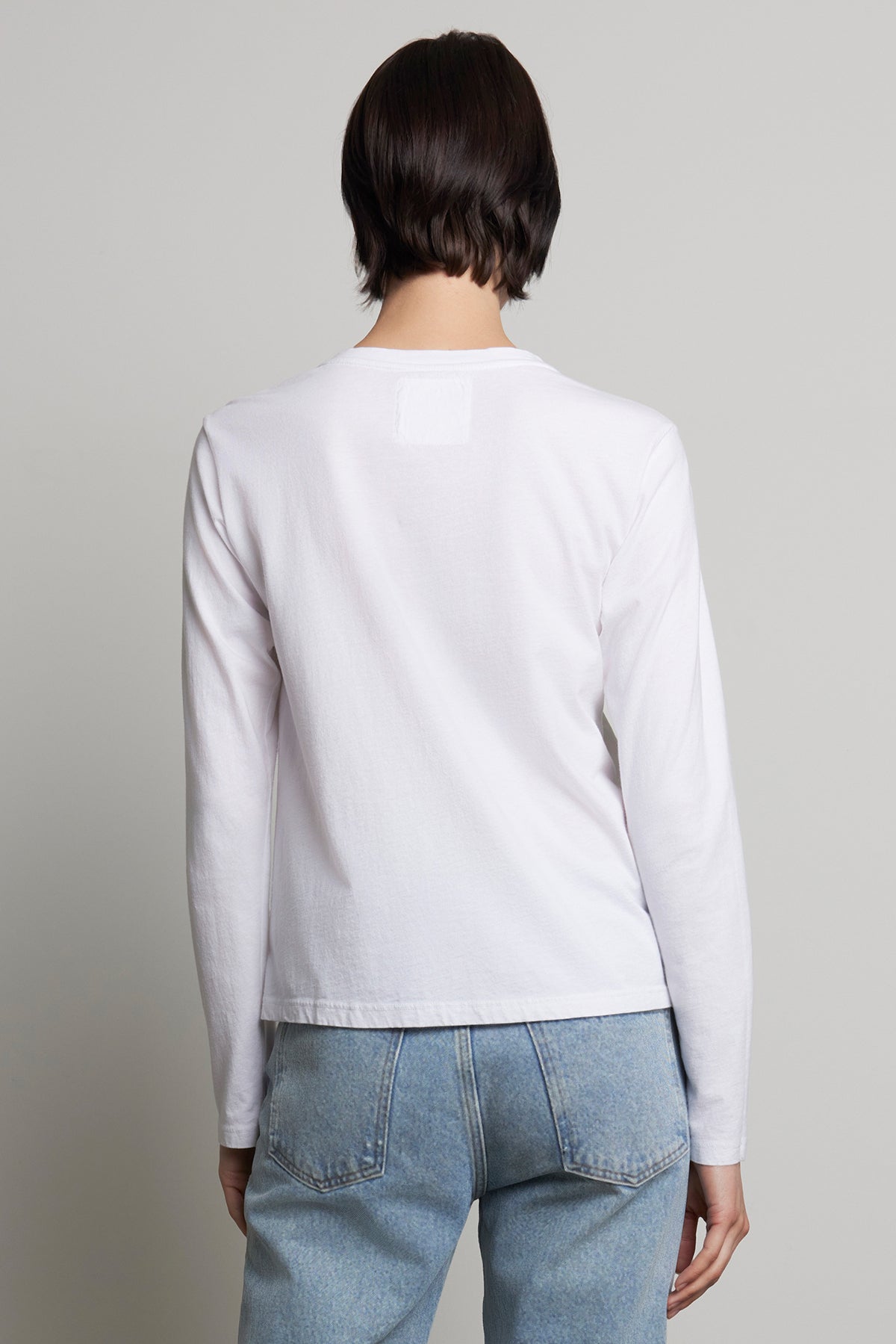   A person is seen wearing the relaxed-fit VICENTE TEE by Velvet by Jenny Graham, a white long sleeve shirt made of organic cotton, paired with blue jeans, and facing away from the camera. 