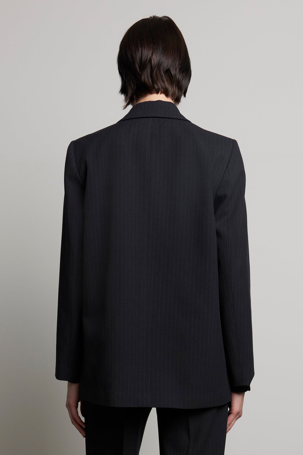   A person with short dark hair is wearing the oversized FRESNO PINSTRIPE BLAZER by Velvet by Jenny Graham, featuring a notched collar, and is facing away from the camera against a plain background. 