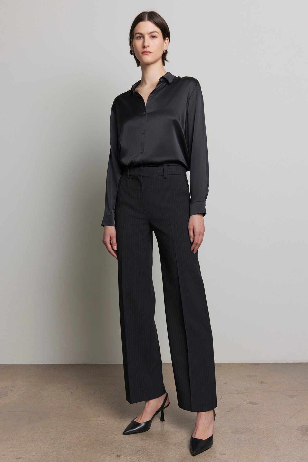   A person stands against a plain background, wearing a dark long-sleeve shirt and the stylish TAHOE PINSTRIPE PANT from Velvet by Jenny Graham, featuring a straight leg and high waist, paired with elegant heels. 