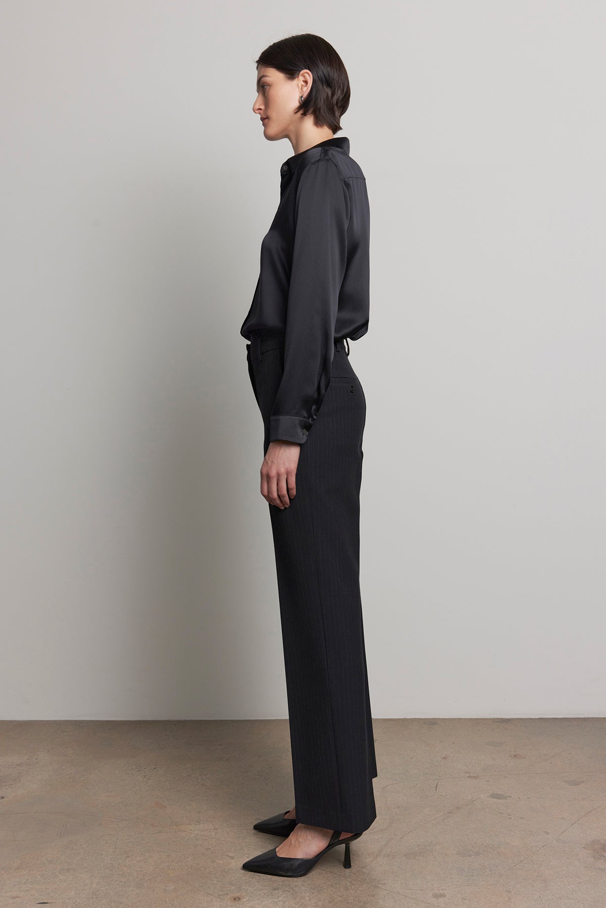 A person standing in profile, wearing a dark long-sleeved shirt paired with the TAHOE PINSTRIPE PANT by Velvet by Jenny Graham and complemented by black heels, against a neutral background.-38318600126657