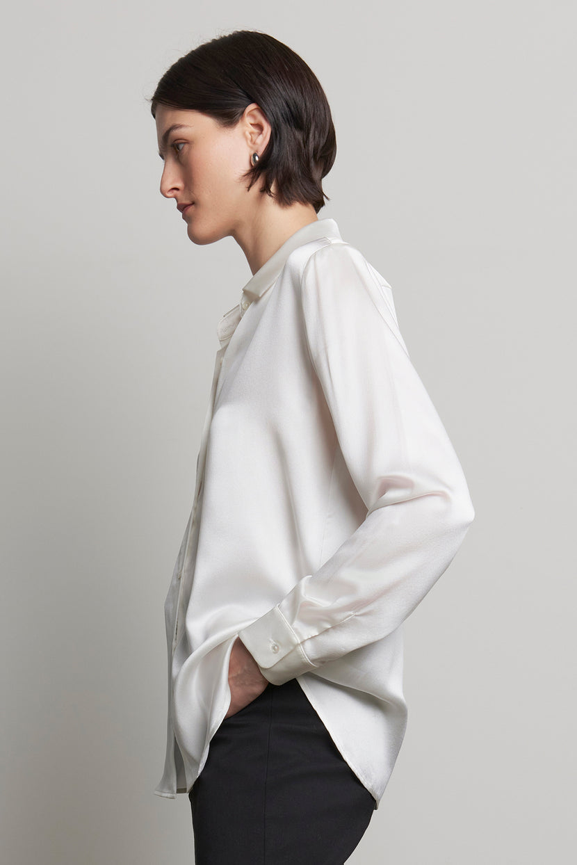 A person wearing the CALABASAS SILK CHARMEUSE BLOUSE by Velvet by Jenny Graham and black pants, with hands in pockets, is facing left against a gray background.