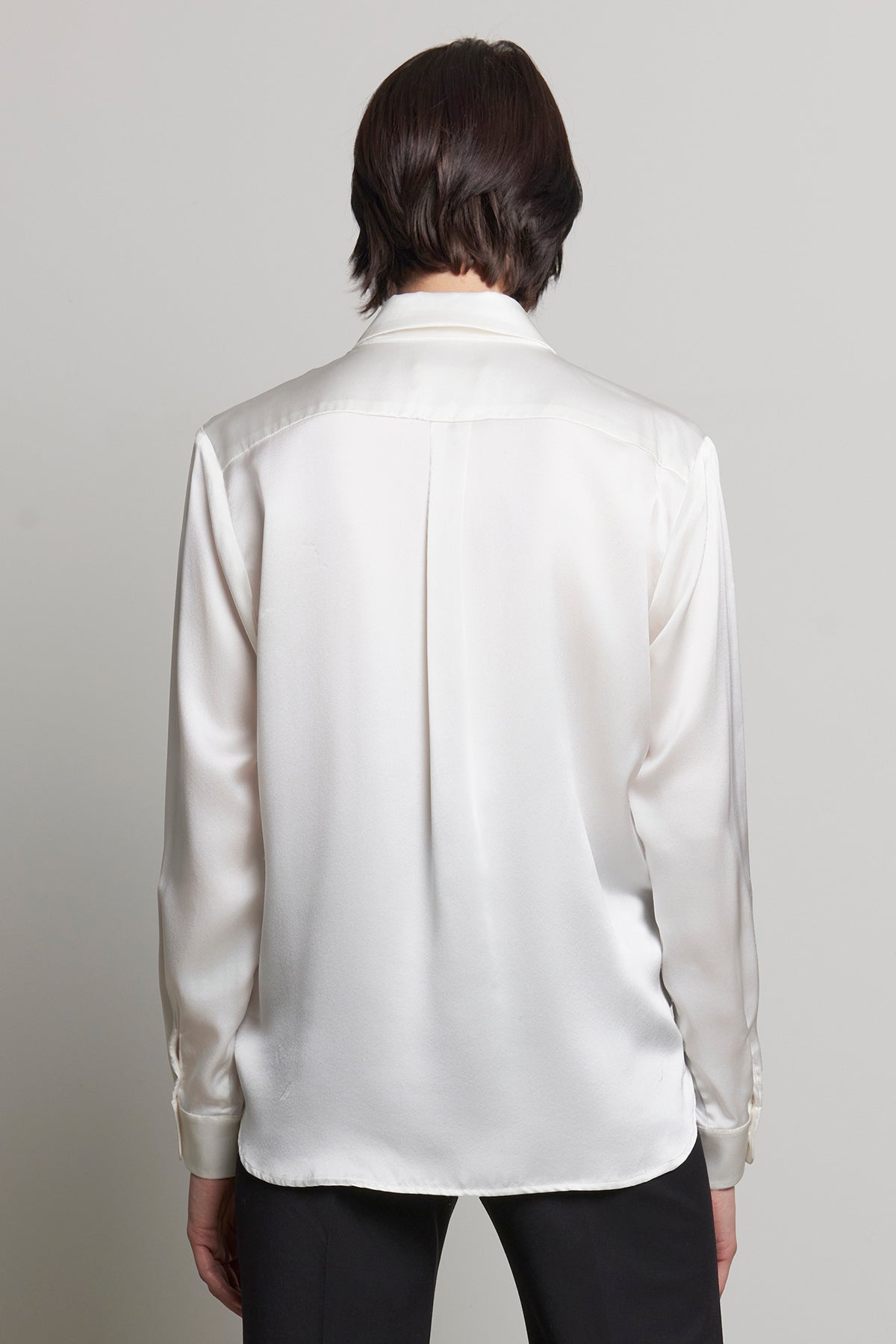   A person with shoulder-length hair stands facing away, wearing a CALABASAS SILK CHARMEUSE BLOUSE by Velvet by Jenny Graham in white with long sleeves. 