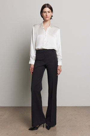 Against a plain background, a person is standing in the MARIPOSA PANT by Velvet by Jenny Graham, featuring a white blouse and black, soft suiting fabric with flared legs.