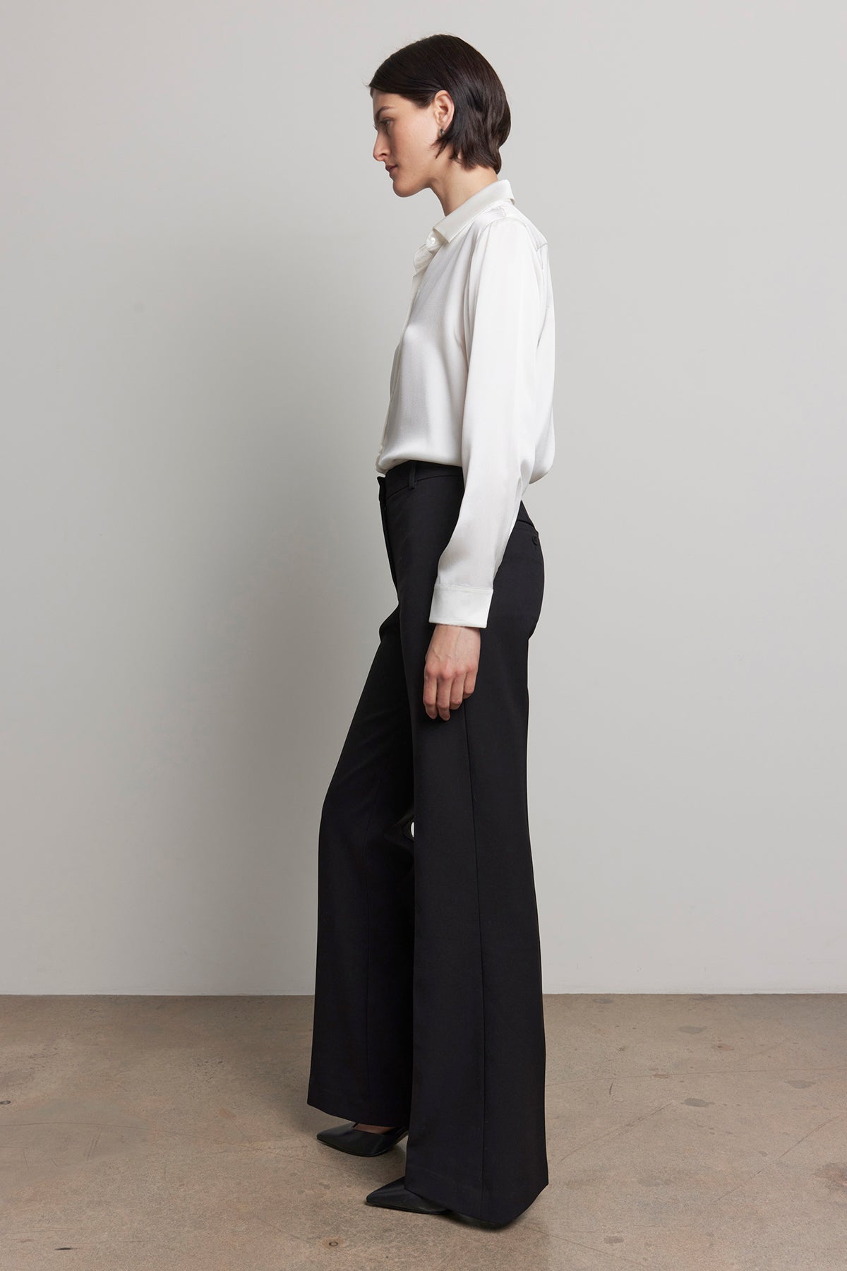 A person stands in profile wearing a white shirt and the MARIPOSA PANT by Velvet by Jenny Graham, featuring black soft suiting fabric with flared legs, against a plain background.-38318589968577