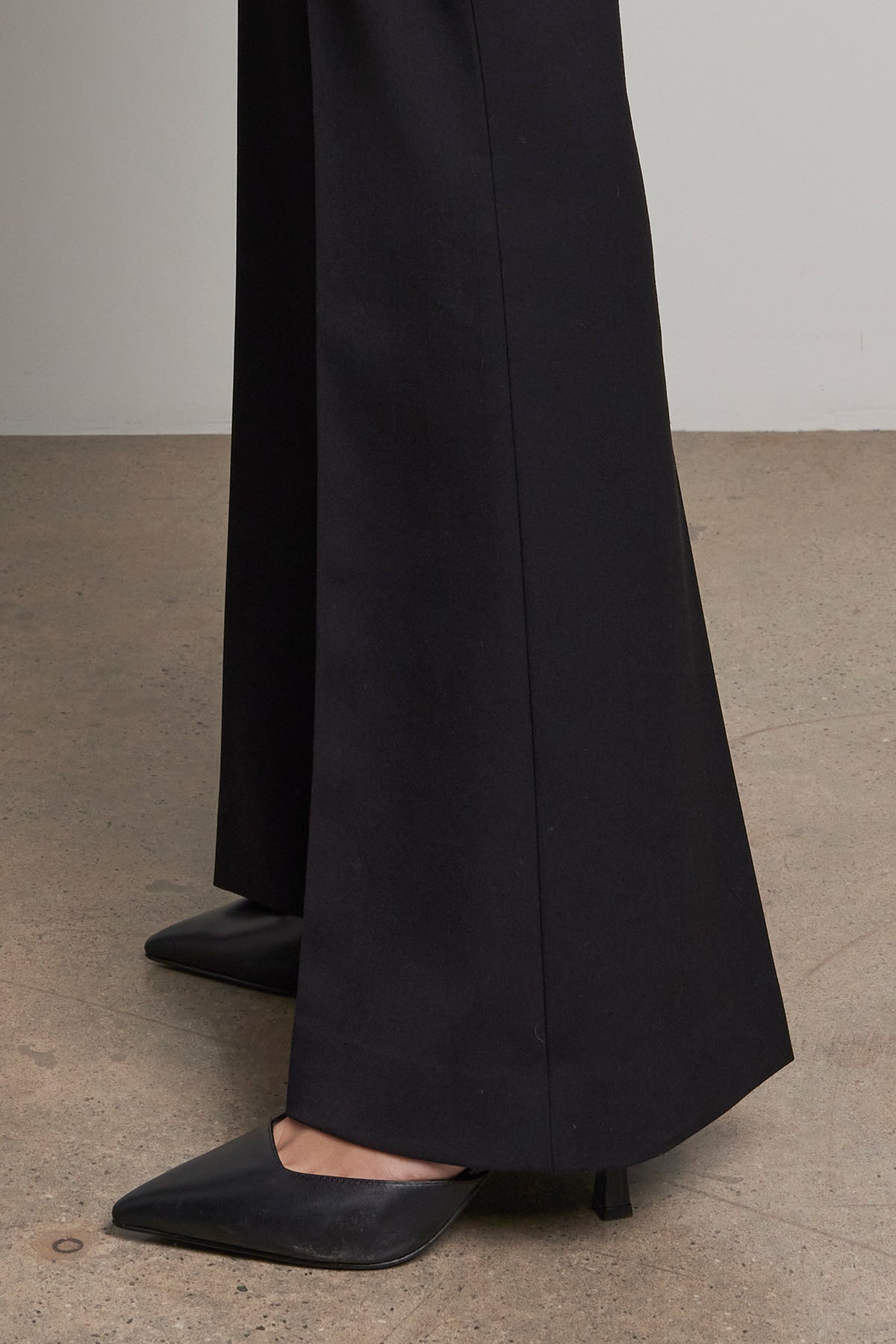 A person stands on a concrete floor wearing the MARIPOSA PANT by Velvet by Jenny Graham, which features black wide-leg pants with a tailored closure, paired with pointed black heels.-38318589903041