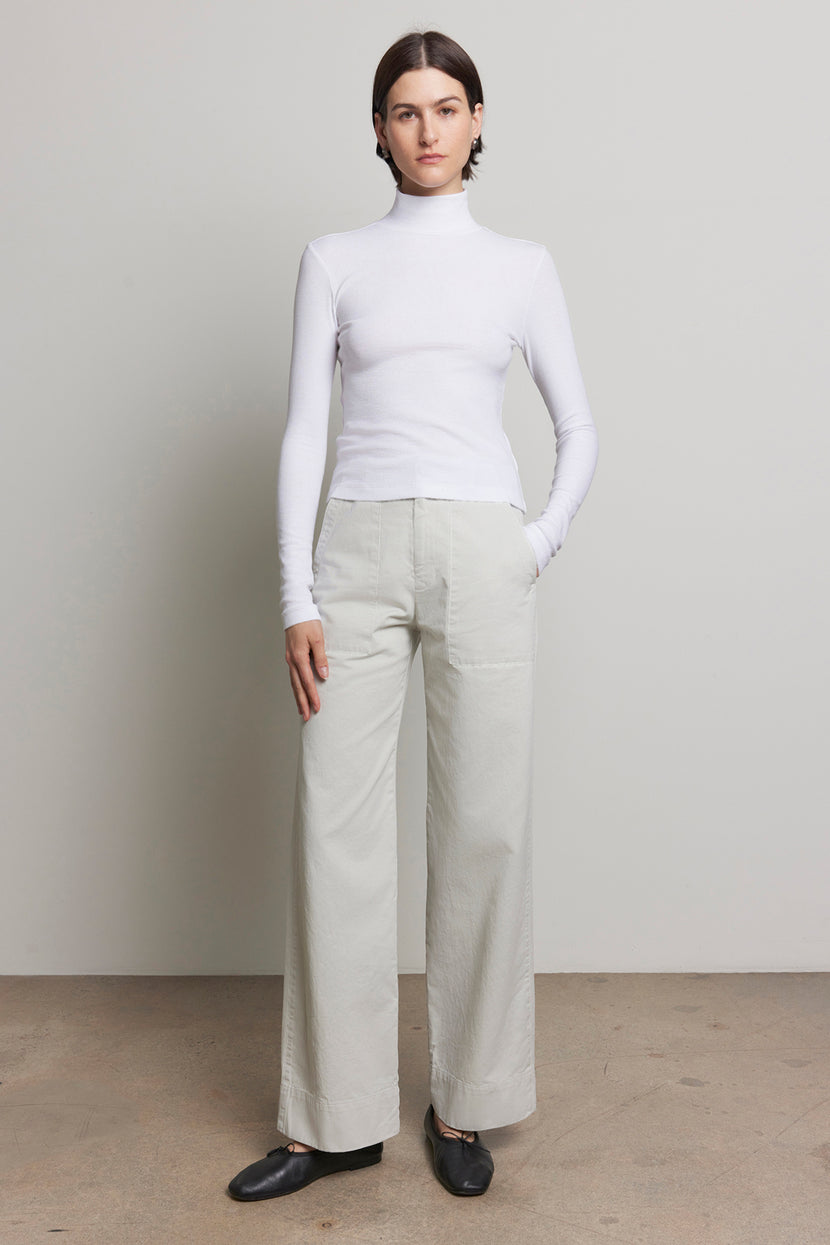 A person standing in a white turtleneck and Velvet by Jenny Graham's VENTURA PANT, which features a utilitarian vibe, with hands in pockets against a neutral background.