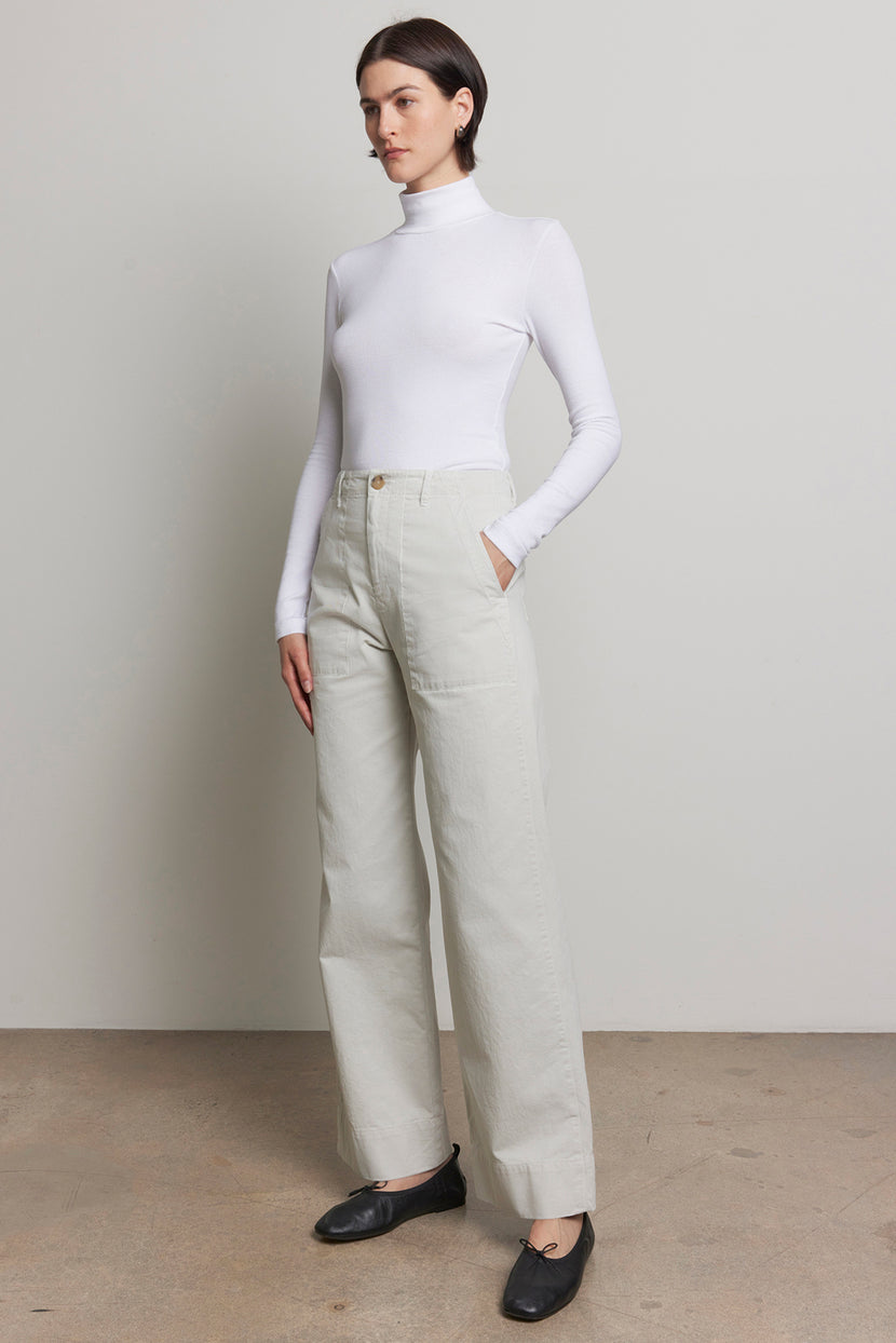 A person stands against a neutral background wearing a white turtleneck, the light-colored VENTURA PANT by Velvet by Jenny Graham, and black shoes.