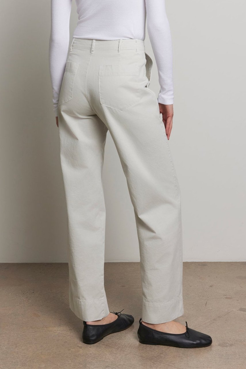   A person stands facing away, donning a white turtleneck and the VENTURA PANT, a pair of light-colored, wide-leg high-waisted pants crafted from cotton twill by Velvet by Jenny Graham. Black shoes complete the utilitarian ensemble against a plain background. 